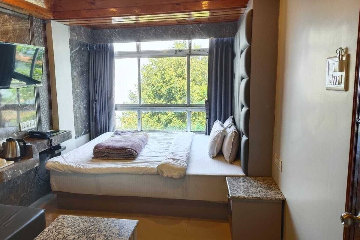 Een slaapkamer met een bed, een aan de muur bevestigde televisie, een raam met gordijnen en een klein tafeltje met een waterkoker.
