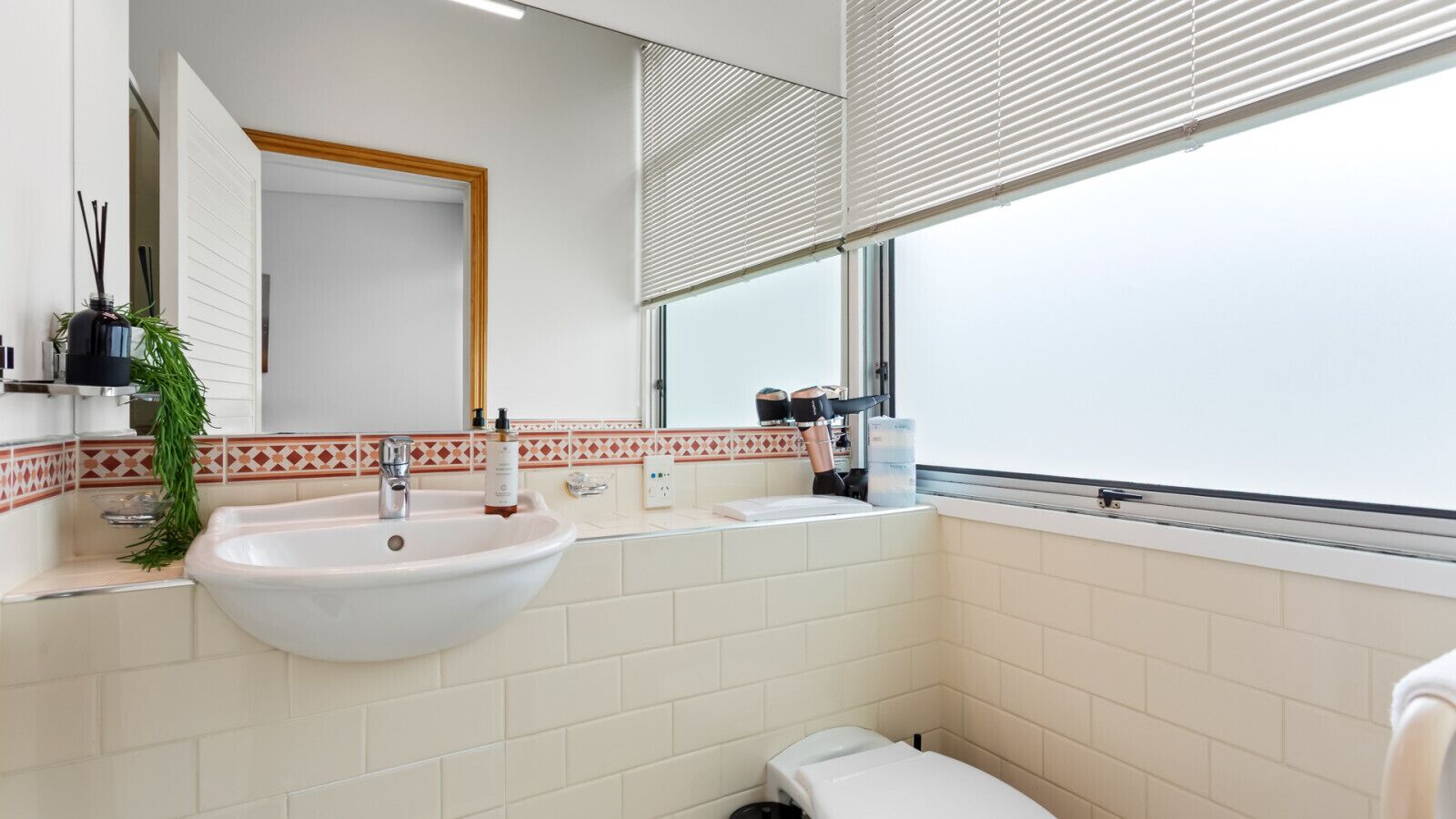 Un baño con un lavamanos blanco, un espejo, una ventana con persianas y un inodoro.