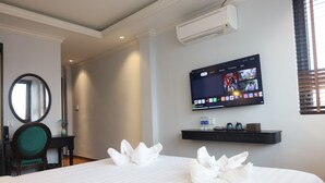 A hotel room with a large bed, a flat-screen TV mounted on the wall, a small shelf with decorative items, and a round mirror on a wooden chair. - Maya Central Hotel & Spa (Hanoi)