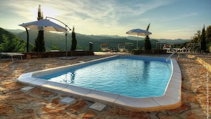 Ein Pool mit Steinböden, zwei Sonnenschirmen und Blick auf Berge.