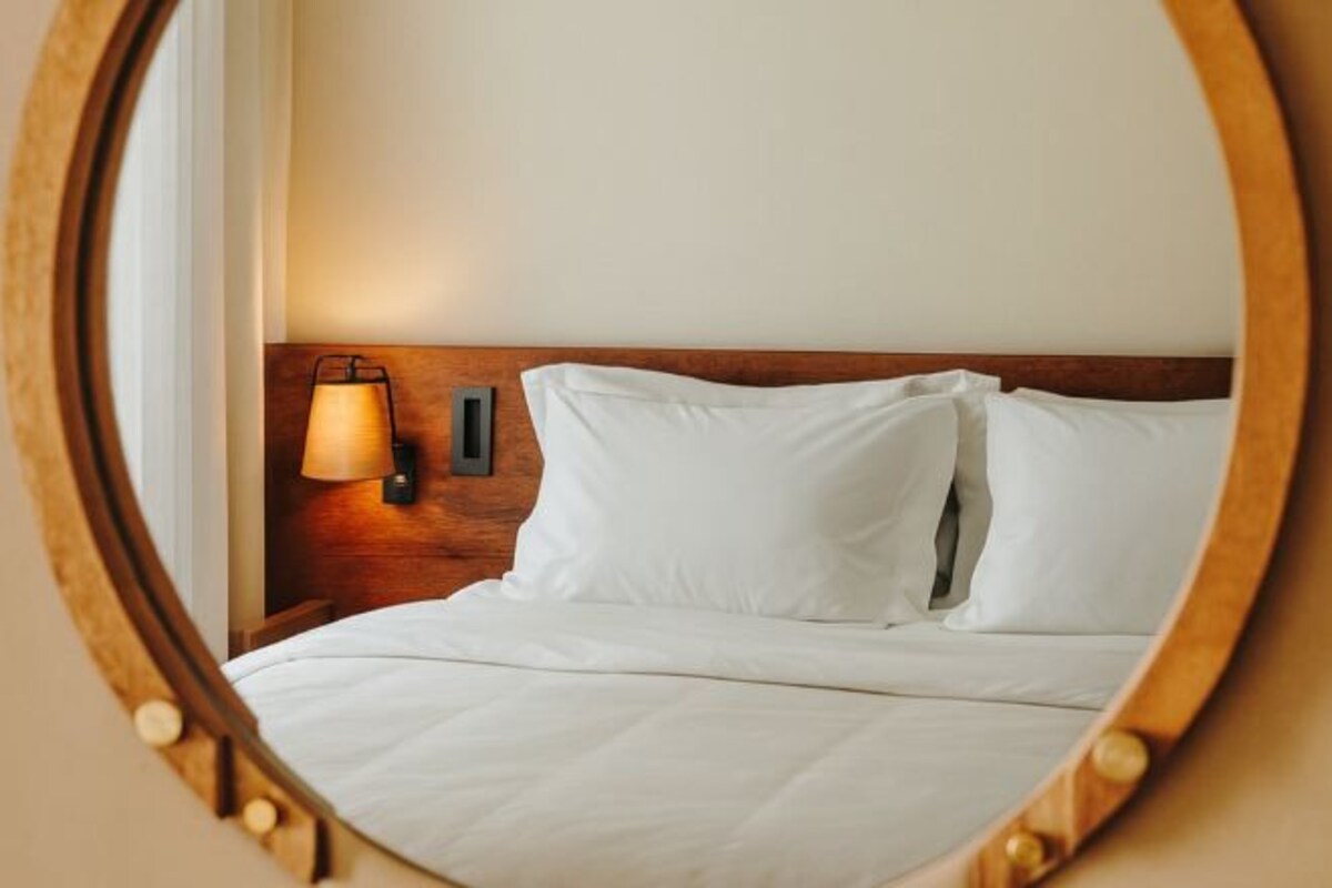 Una cama bien tendida con sábanas blancas, cabecera de madera y lámpara de pared.