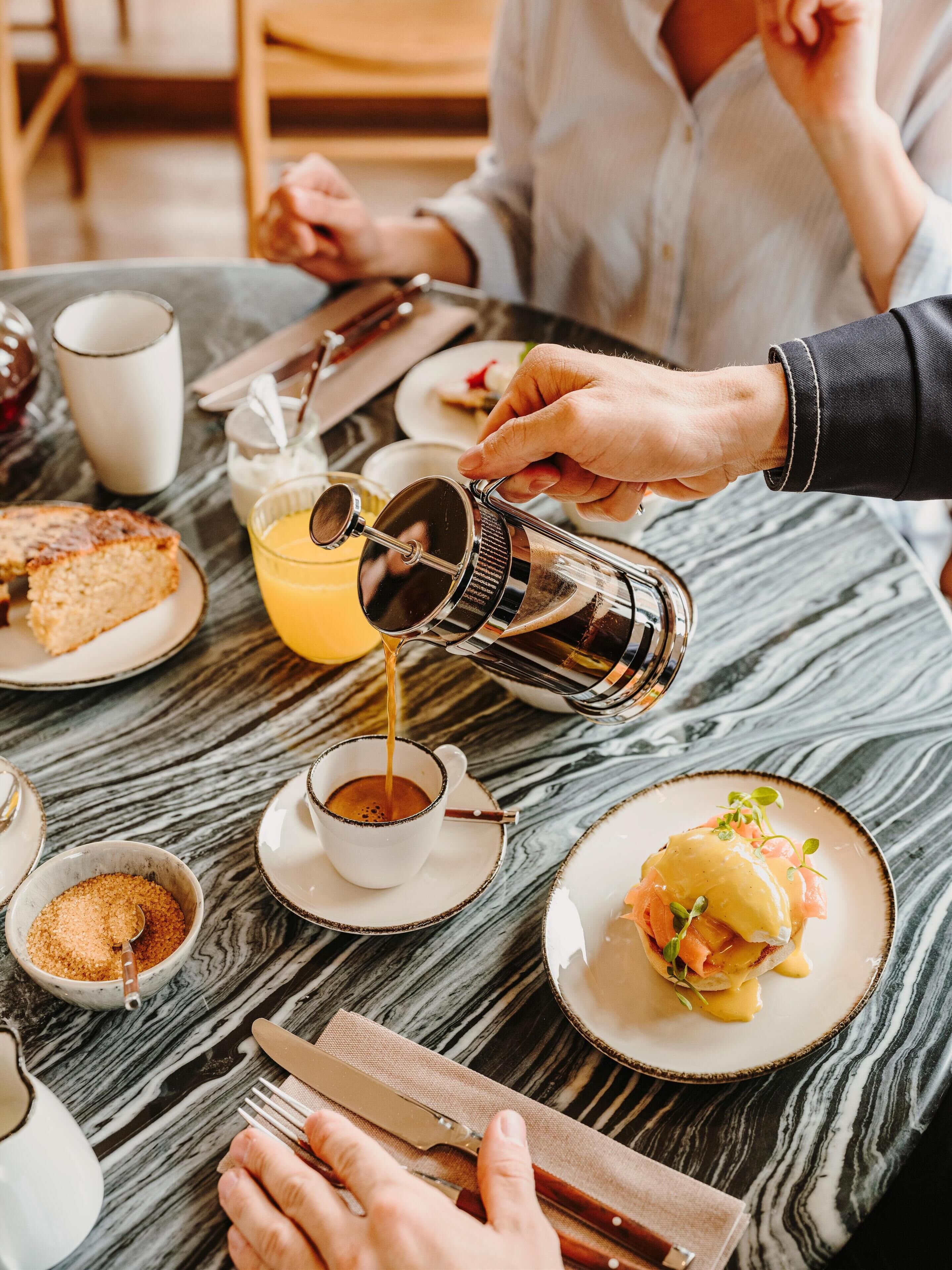 Un desayuno con una cafetera vertiendo en una taza, un plato de comida y un vaso de jugo de naranja sobre una mesa de mármol.