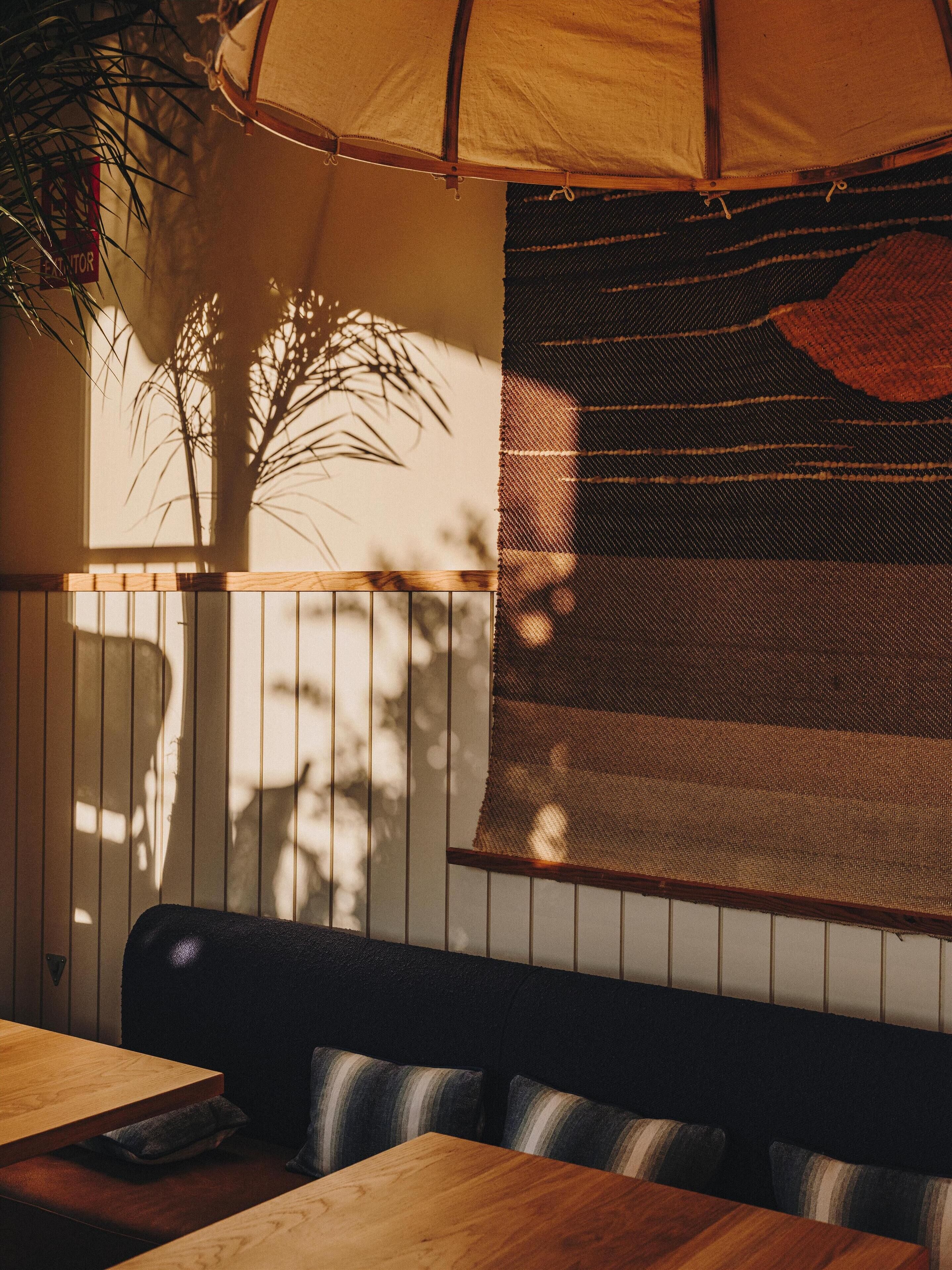 Interior de un restaurante con mesas y bancos de madera, un separador tejido grande y vista a una palmera fuera.