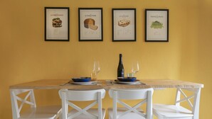 Uma mesa de jantar com quatro ilustrações emolduradas de comida, dois copos de vinho e cadeiras brancas.