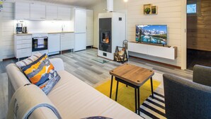 Una sala de estar moderna con sofá, mesa de centro y chimenea. Hay una zona de cocina con armarios blancos y una estufa. Un televisor está montado en la pared sobre la chimenea.