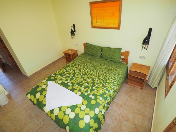 A bedroom with a green polka dot bedspread, a wooden headboard, a nightstand, a wall clock, and a painting above the bed. - Shams Hotel and Dive Centre (Dahab)