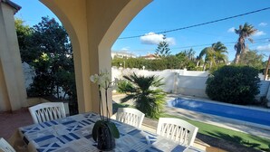 Um alpendre coberto com uma mesa posta para dois, um vaso de planta e vista para uma piscina e vegetação.