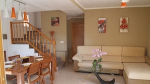 Uma sala de estar com um sofá bege, uma mesa de centro de vidro com uma orquídea rosa, uma mesa de jantar de madeira com cadeiras e uma escada com corrimãos de madeira.