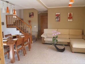 A living room with a beige sofa, a glass coffee table with a pink orchid, a wooden dining table with chairs, and a staircase with wooden railings. - Villa Naomi (Les Tres Cales)
