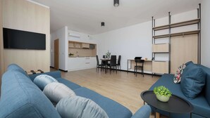 Ein modernes Wohnzimmer mit einem blauen Sofa, einem kleinen Tisch mit Pflanze und einer Küche im Hintergrund.