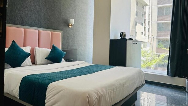 A hotel room with a bed, a nightstand, a window with a view of buildings, and a door. - HOTEL SAWARIYA (Ahmedabad)