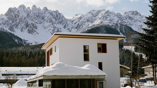 Ein modernes, zweistöckiges Gebäude mit flachem Dach, umgeben von schneebedeckten Bergen und Bäumen.