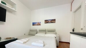 Un letto rifatto con lenzuola bianche e un asciugamano decorativo, un comodino con un piatto di cibo e due quadri appesi al muro.