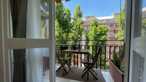 Un balcone con tavolo e sedie, vista su edifici e alberi, e cielo sereno.