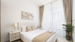A modern bedroom with a large bed, bedside tables, a nightstand, a potted plant, and a window with sheer curtains. - Close to Community Park, Minutes Walk from Circle Mall, and Short Drive to Dubai Miracle Garden (Dubai)