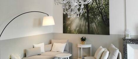 Un salon moderne avec un canapé gris, un canapé blanc plus petit, une table ronde blanche, une lampe blanche, un lustre et un grand tableau sur le thème de la forêt accroché au mur.