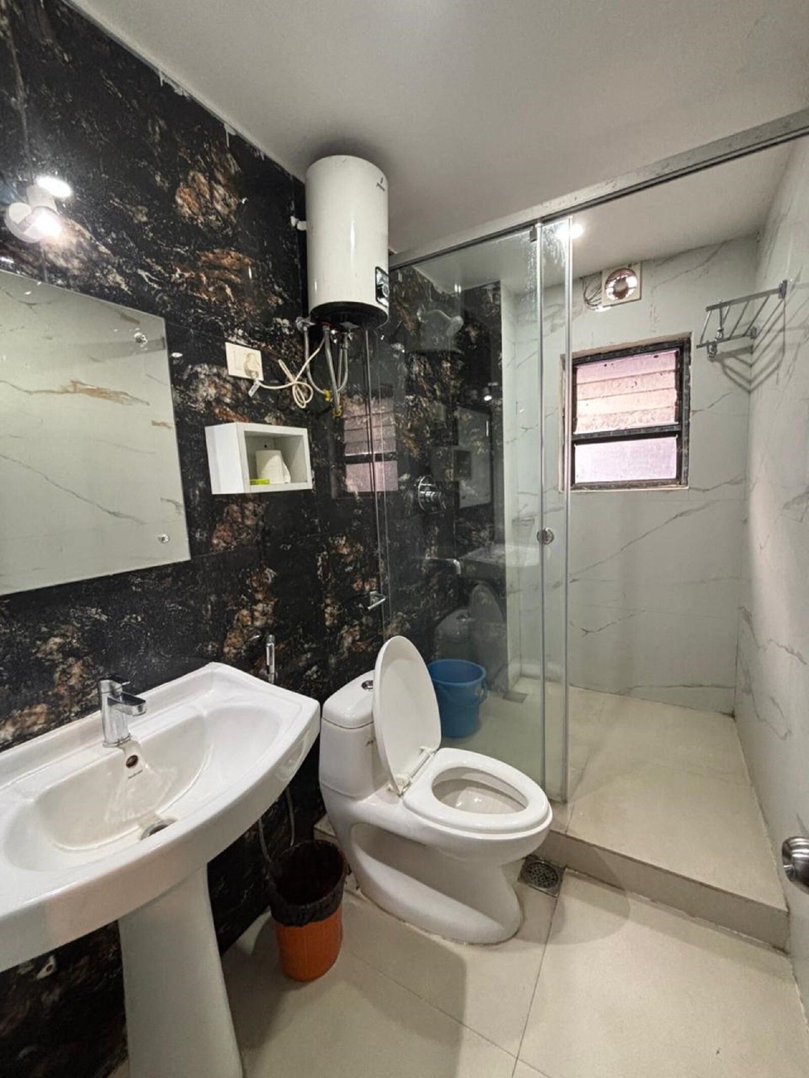 Casa de banho com parede de mármore, lavatório, sanita e chuveiro, além de uma pequena janela.