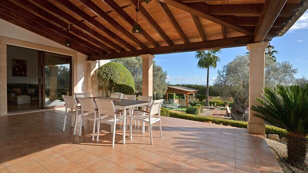 Un patio exterior con una pérgola de madera, una mesa de comedor con sillas y un jardín bien cuidado con una palmera y un pabellón.
