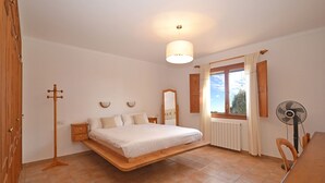 Un dormitorio con cama nido, ventilador de pie, ventana con cortinas, radiador y una mesa de madera.