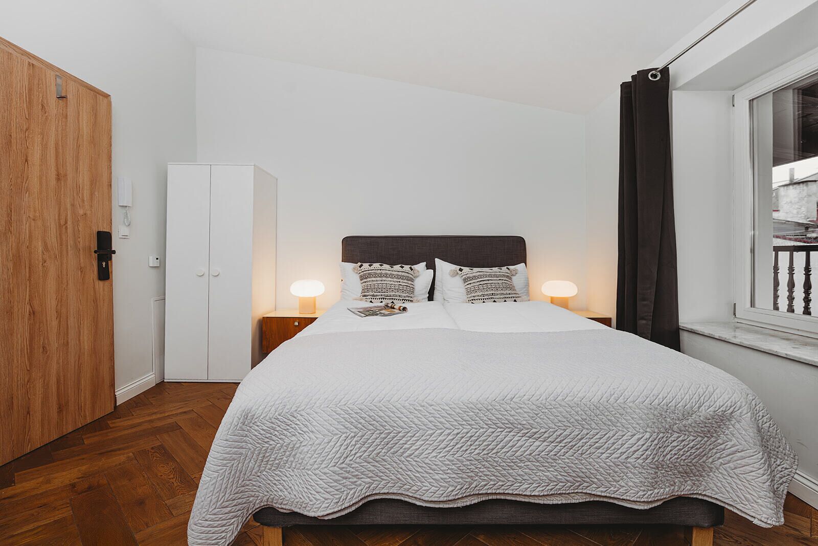 A neatly arranged bedroom with a bed, bedside lamps, a wardrobe, and a window with curtains.
