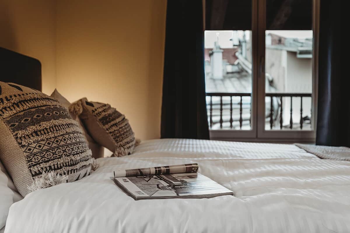 Ein ordentlich bezogenes Bett mit gemusterten Kissen, ein Buch und Blick auf Gebäude durch ein Fenster.