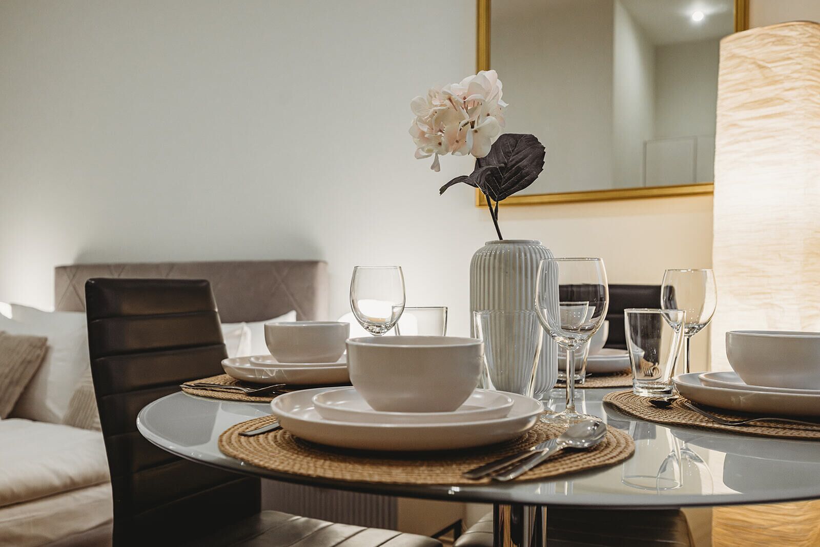 Une table à manger dressée avec de l’assiette blanche, des bols et des verres à vin, un vase rempli de fleurs et une lampe.