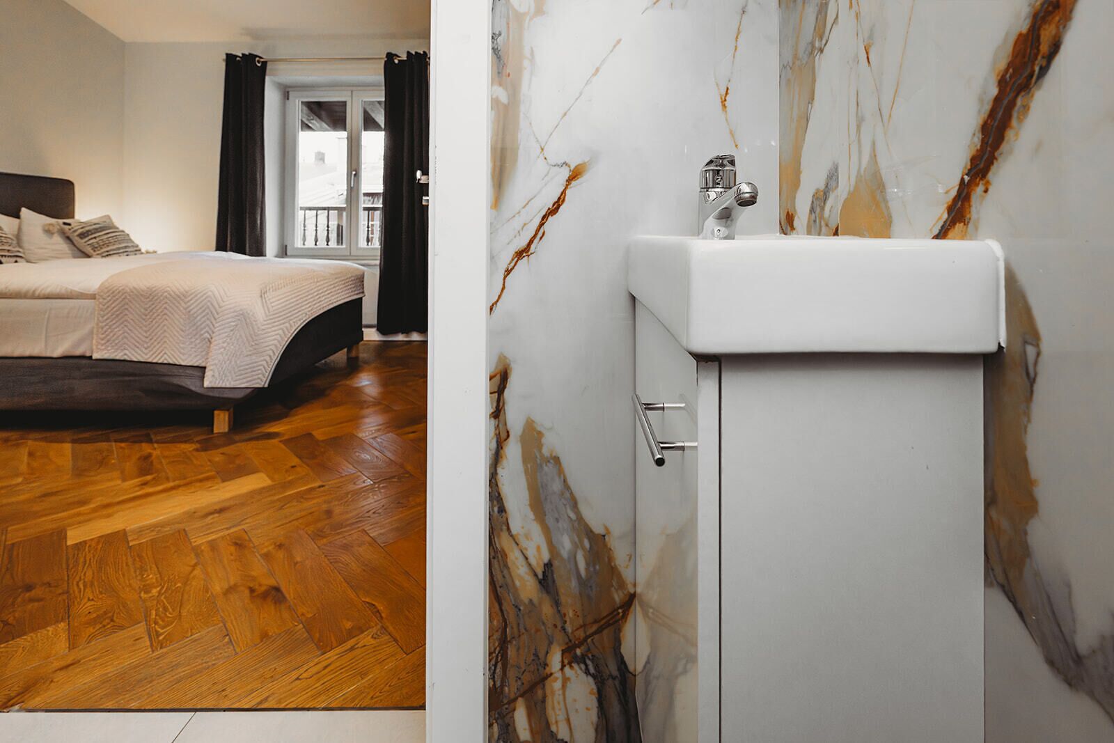 A bedroom with a wooden floor, a bed with a headboard, and a bathroom with a marble wall and a sink.