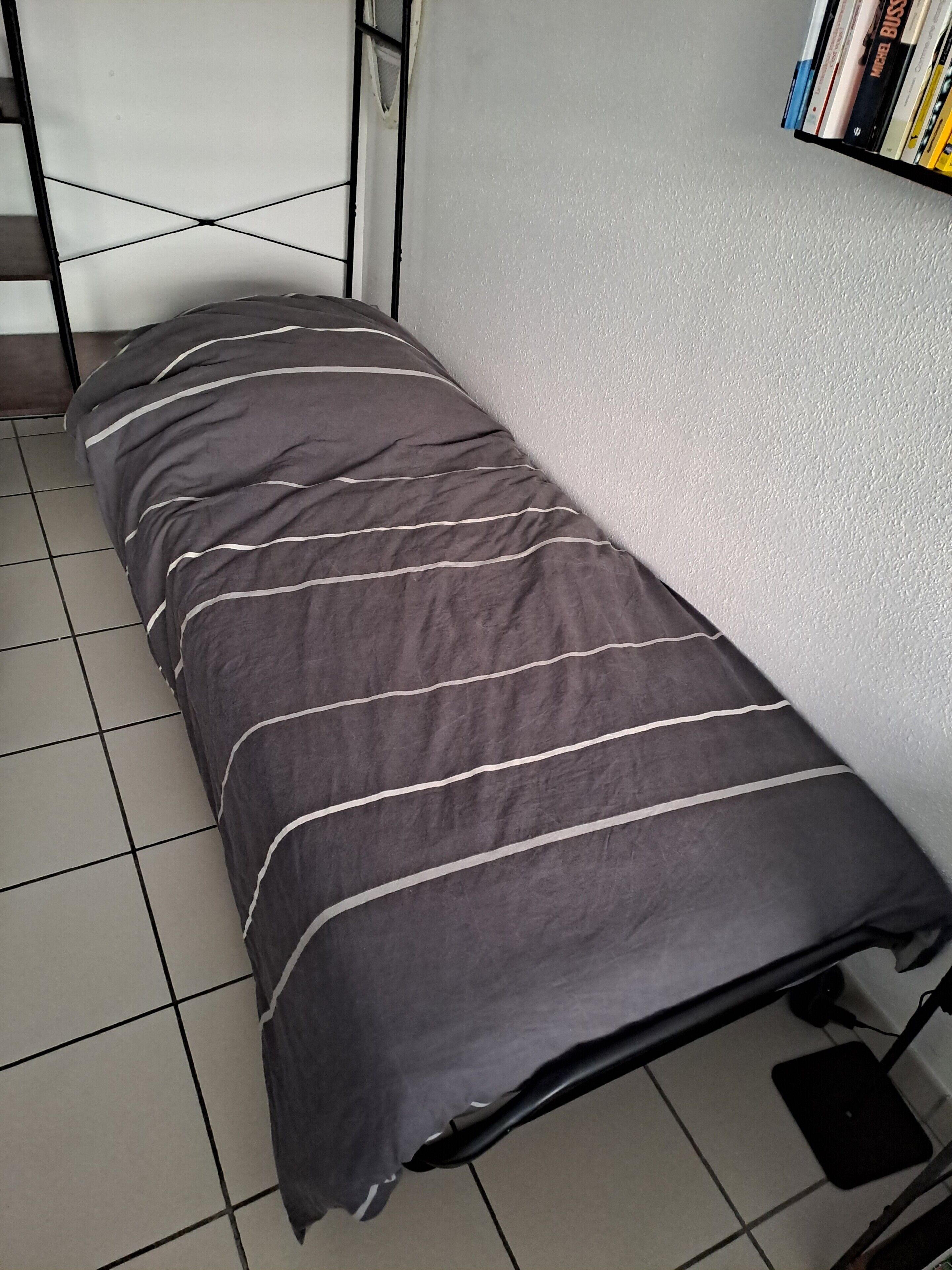 En liten säng med ett randigt täcke, en bokhylla full med böcker och ett klinkergolv.