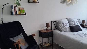 Et soveværelse med seng, gyngestol, natbord, lampe, væghylle med bøger og en plante, samt en dekorativ vægdekoration.
