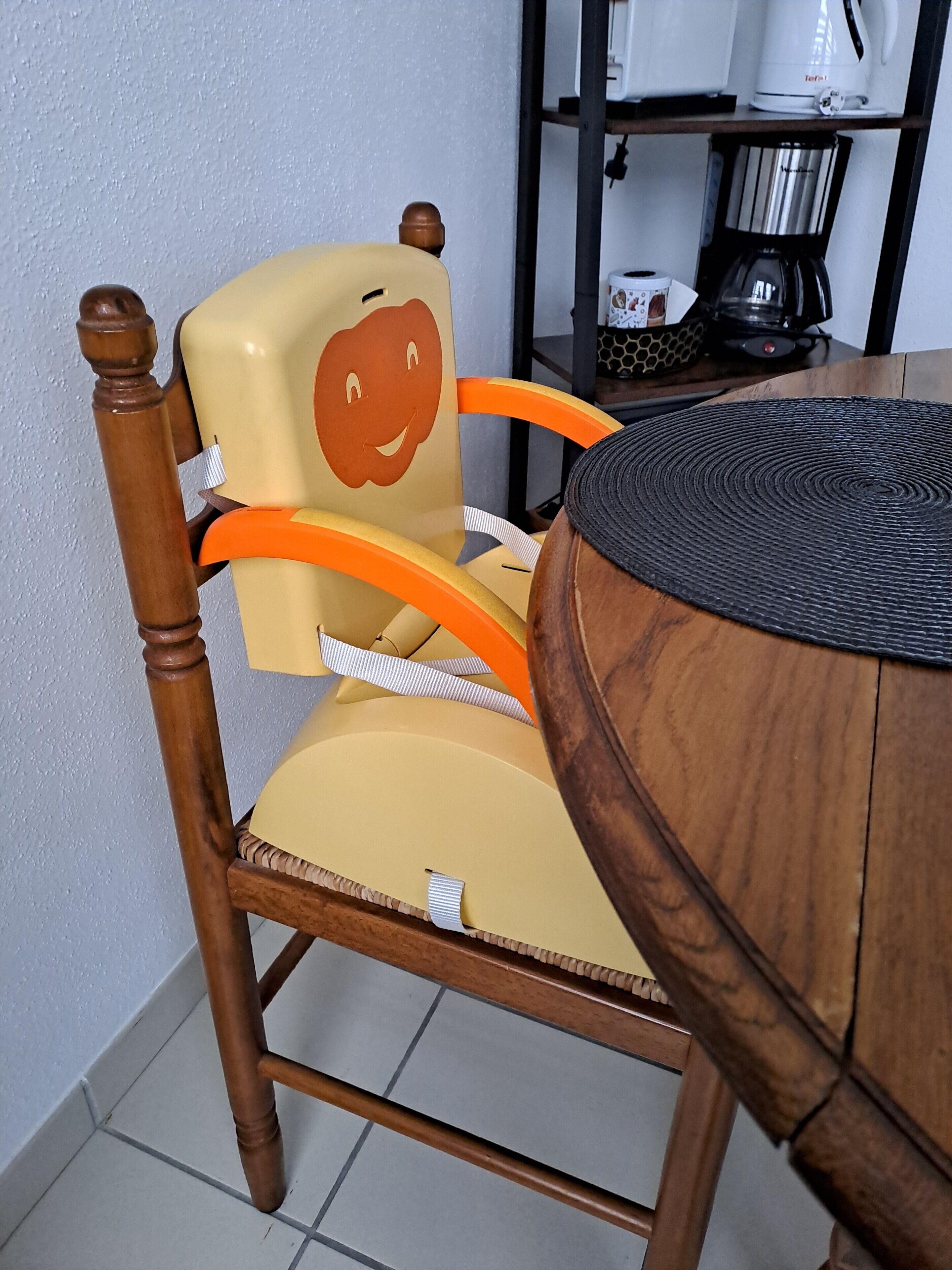 A wooden chair with a cushion and a face design, a round table, and a coffee maker on a shelf.