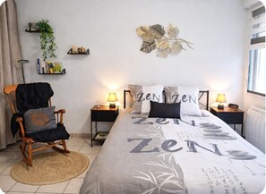 A bedroom with a bed, two bedside tables, a chair, a floor lamp, a wall shelf with books and a plant, and a decorative leaf artwork.