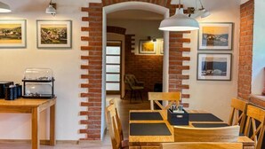 Un comedor con arcos de ladrillo, mesas y sillas de madera. Hay cuadros en las paredes y una cafetera sobre la mesa.