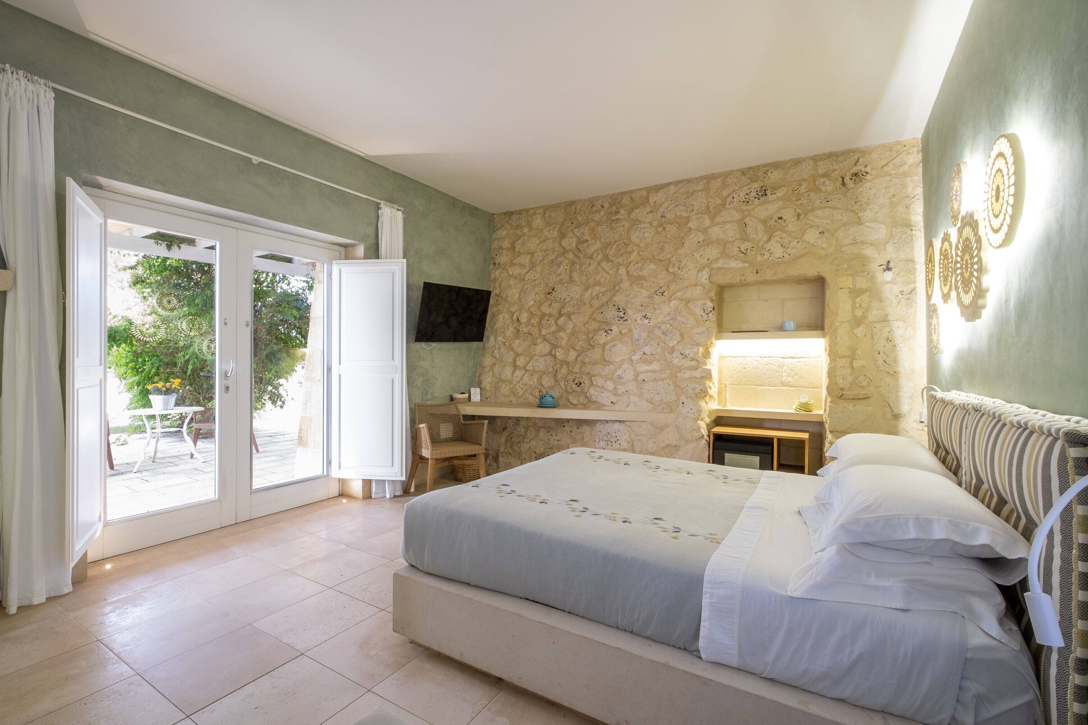A bedroom with a bed, a desk, a chair, a stone wall, and a TV.
