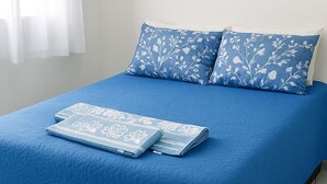 Una cama con colcha azul y almohadas a juego, una manta doblada y una persiana blanca.
