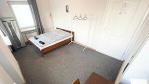 Ein kleines Zimmer mit einem Einzelbett, einem Holzrahmen, einem grauen Teppich, einem Fenster mit Vorhängen, einem Heizkörper, einer weißen Tür und einem Kleiderständer mit Kleiderbügeln.