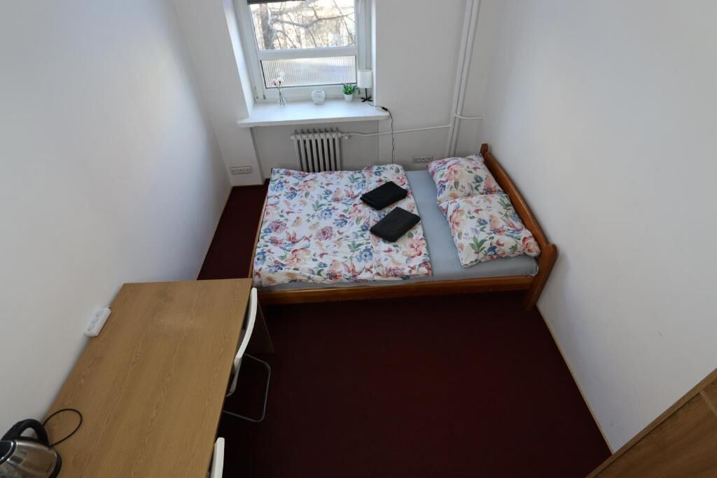 Een kleine kamer met een enkel bed, een houten bedframe, bloemenbeddengoed, een bureau met stoel, en een radiator onder een raam.