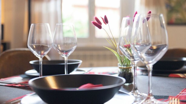 Une table à manger dressée avec des verres à vin, un bol noir et un petit vase avec des fleurs.