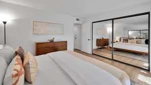 A modern bedroom with a large bed, a wooden dresser, a mirror, and a sliding door leading to another room. - Sunny Desert Escape - A Ryson Property (Palm Springs)