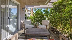 A covered patio with two white cushioned chairs and a wooden table, surrounded by lush greenery and a view of palm trees. - Sunny Desert Escape - A Ryson Property (Palm Springs)