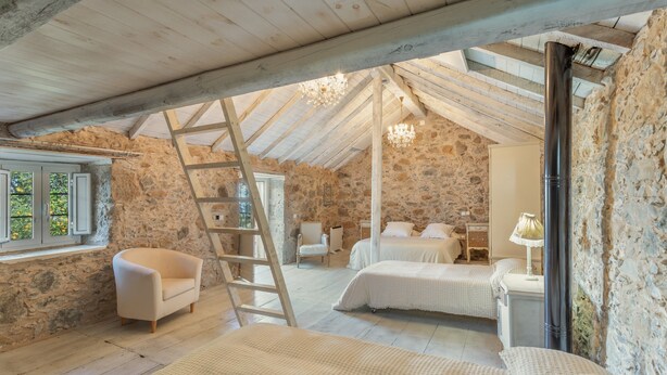 Un dormitorio rústico con paredes de piedra, vigas de madera, una escalera, una silla y dos camas.
