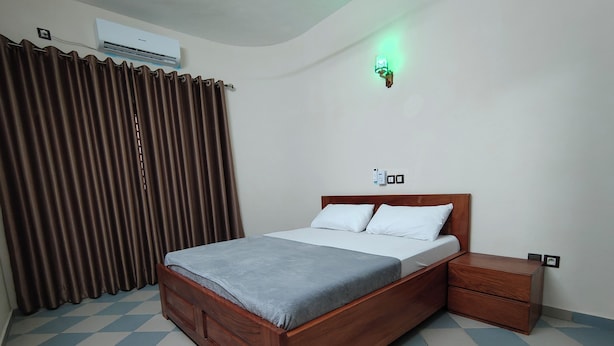 A neatly made bed with a grey blanket, a wooden nightstand, a wall-mounted light, and a window with brown curtains. - Room-executive Queen Size Room In Cotono (Cotonou)