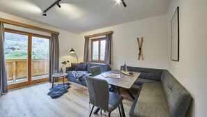 Ein modernes Wohnzimmer mit Sofa, Esstisch und Stühlen. Große Fenster mit Vorhängen, eine Holzterrasse draußen und ein Skiständer an der Wand.