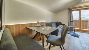 Une pièce chaleureuse avec une table à manger, deux canapés gris, un mur décoratif en bois et une grande fenêtre donnant sur l’extérieur.