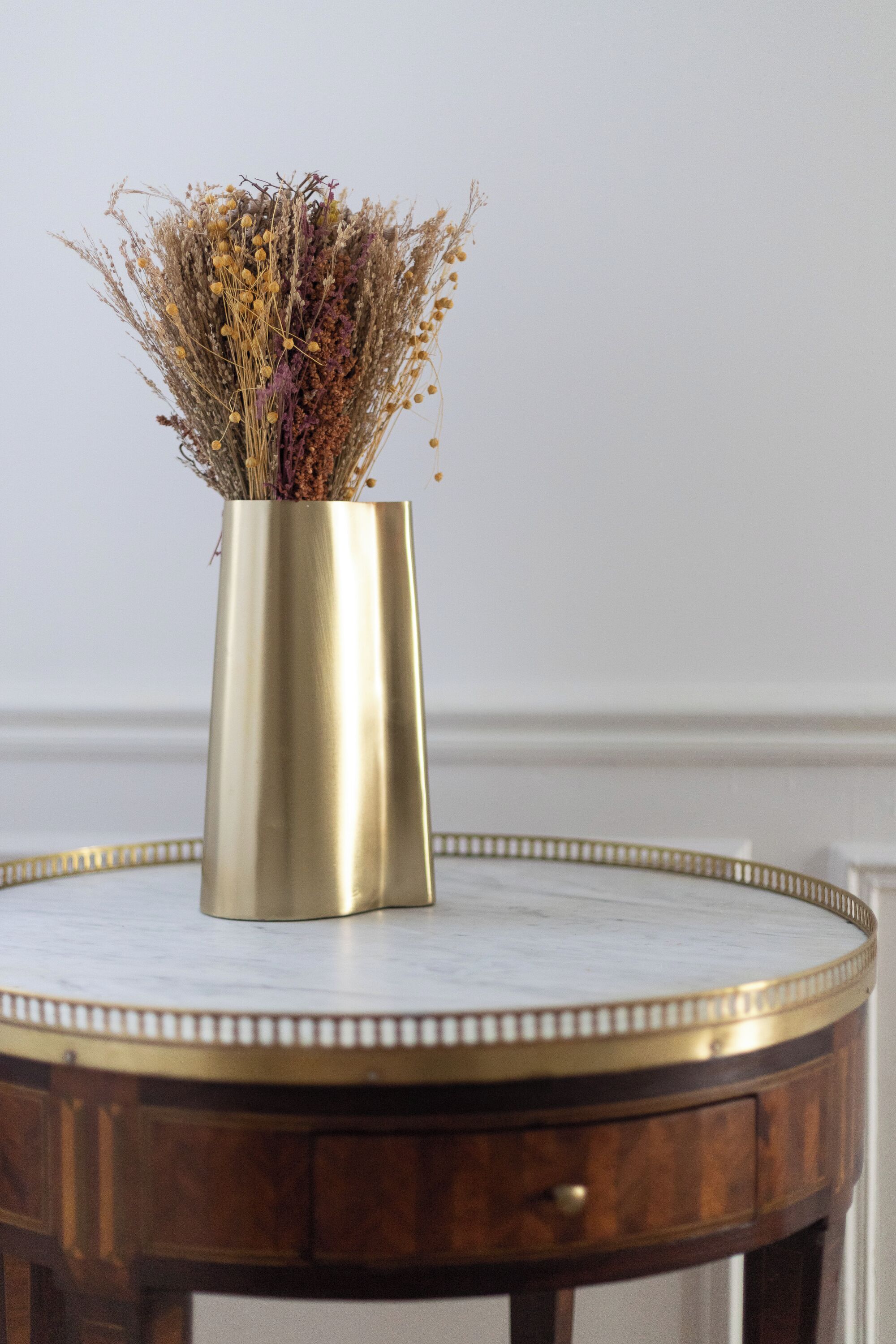 Een ronde houten tafel met een gouden vaas gevuld met gedroogde bloemen.