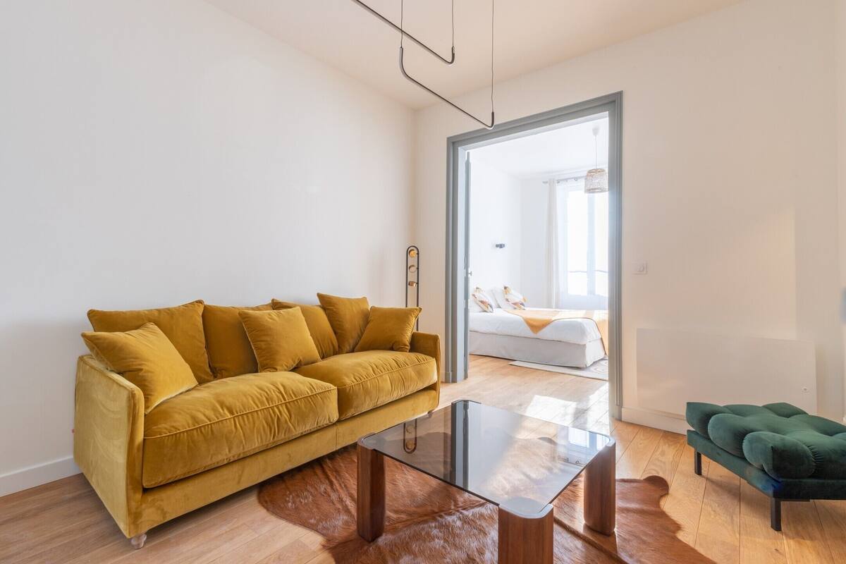 A modern living room with a mustard-colored sofa, a glass coffee table, and a green ottoman.