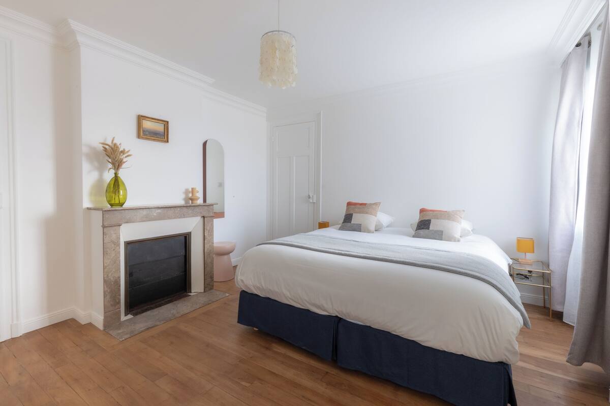 A bedroom with a bed, a fireplace, a nightstand, and a mirror on the wall.