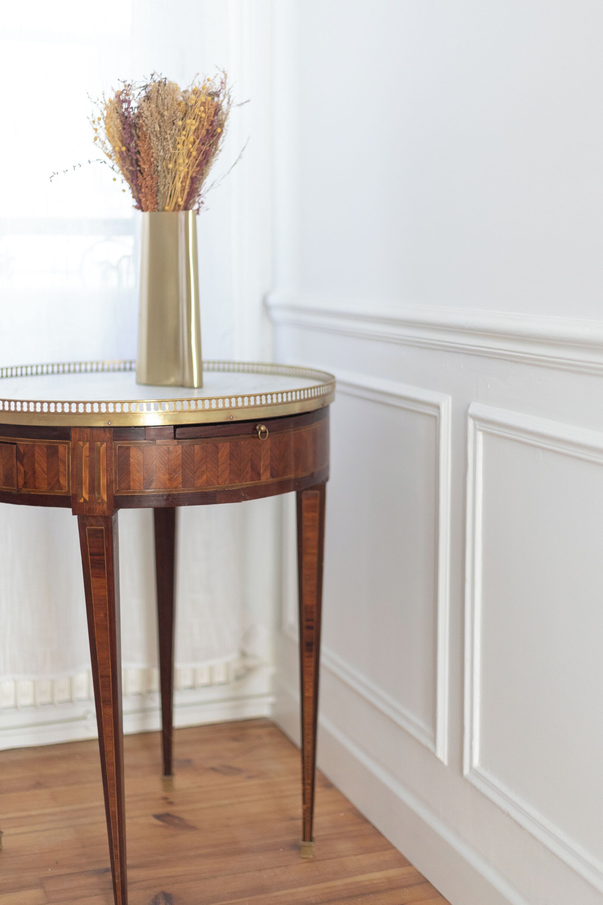 A round wooden table with a gold-colored vase containing dried flowers, placed against a white wall with decorative molding.