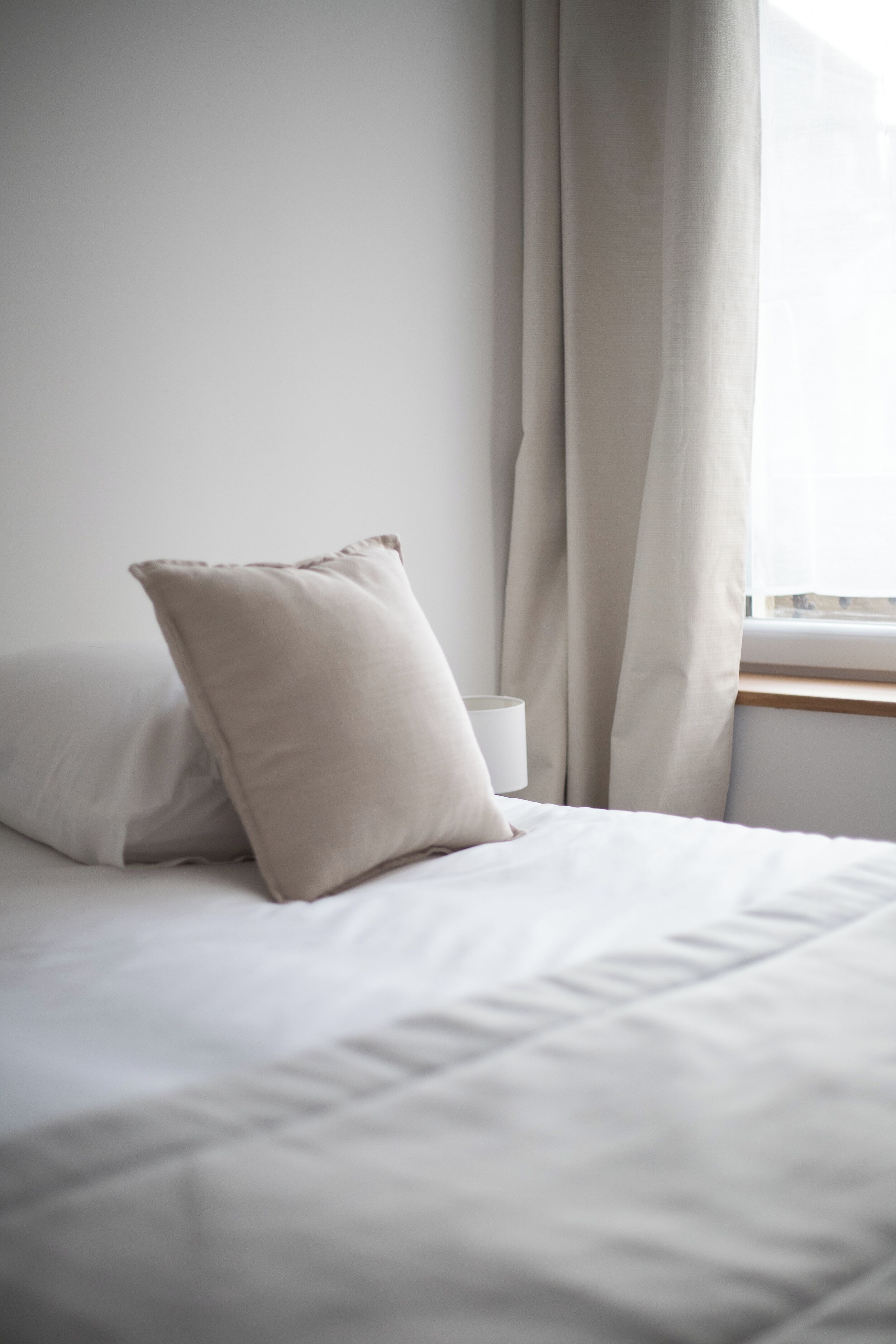A neatly made bed with white linens and two pillows, a bedside table with a white lamp, and a window with sheer curtains.