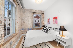 A bedroom with a brick wall, a large window, a bed with white bedding, black and white pillows, a bedside table with a lamp, and two framed artworks on the wall.