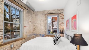 Un dormitorio con paredes de ladrillo visto, un ventanal grande, una cama con colcha blanca, una lámpara negra y una obra de arte en la pared.